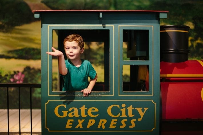 Young boy in a teal shirt leaning out of a train exhibit at the Miriam P. Brenner Children's Museum.