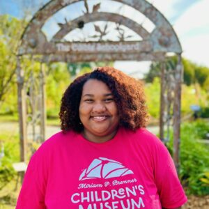 Smiling woman in pink miriam p. Brenner children's museum t-shirt outdoors.